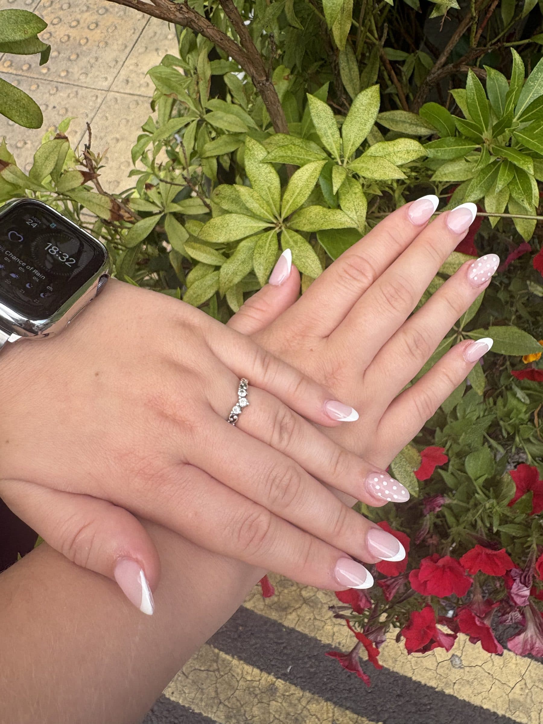 Two hands with almond white French tips, one nail on each hand finished with small white polka dots, laid over green leaves.